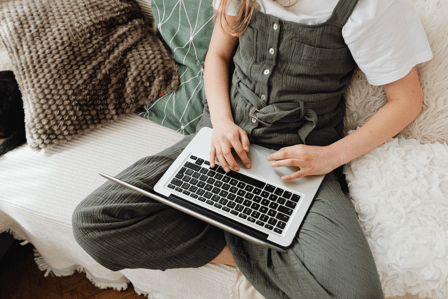 Young white person sits on a white couch, their face out of view. They are typing on a silver laptop.