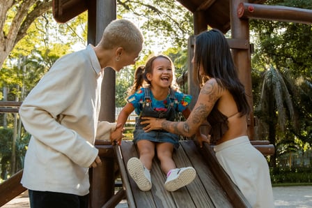 A young lesbian couple playfully help a little girl as she comes down a slide. They are all smiling.