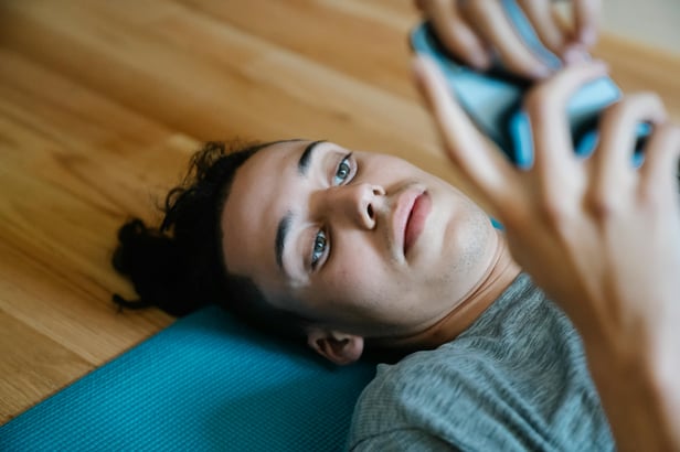 White teen with dark curly lies on yoga mat on timber floor. They are looking blankly at the smart phone in their hands.